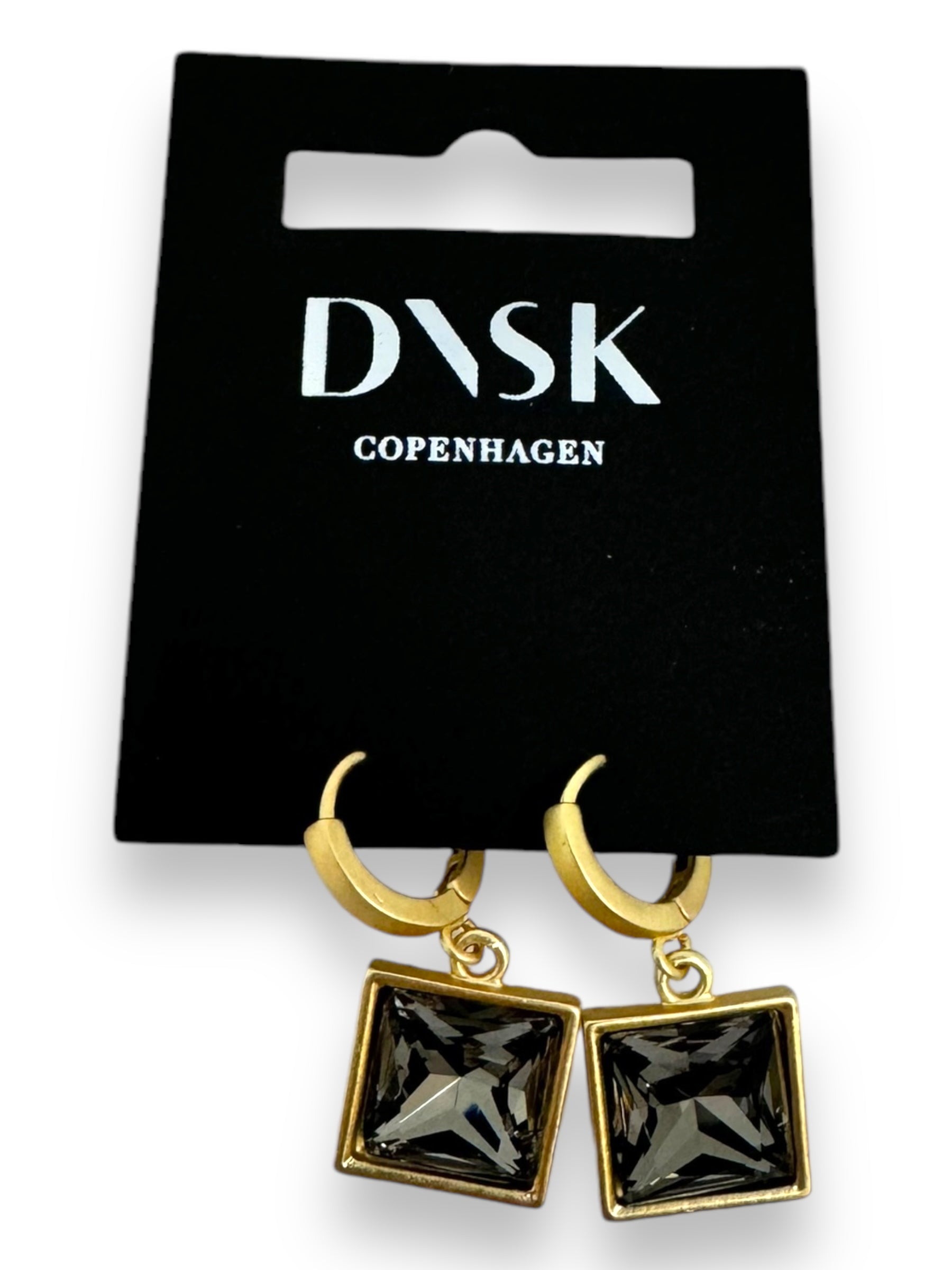 The Audrey Black Diamond Gold Plated earrings by DNSK Copenhagen feature a minimalist Nordic design with gold square hoops and dark gem pendants, crafted from high-quality brass. They are elegantly presented on a sleek black display card, offering versatile jewelry that effortlessly complements any style.