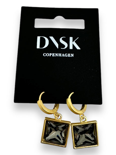 The Audrey Black Diamond Gold Plated earrings by DANSK Copenhagen feature gold hoops with square black gemstone pendants in a Nordic design, displayed on a black card with the brands text. These minimalist jewelry pieces are versatile accessories for any occasion.