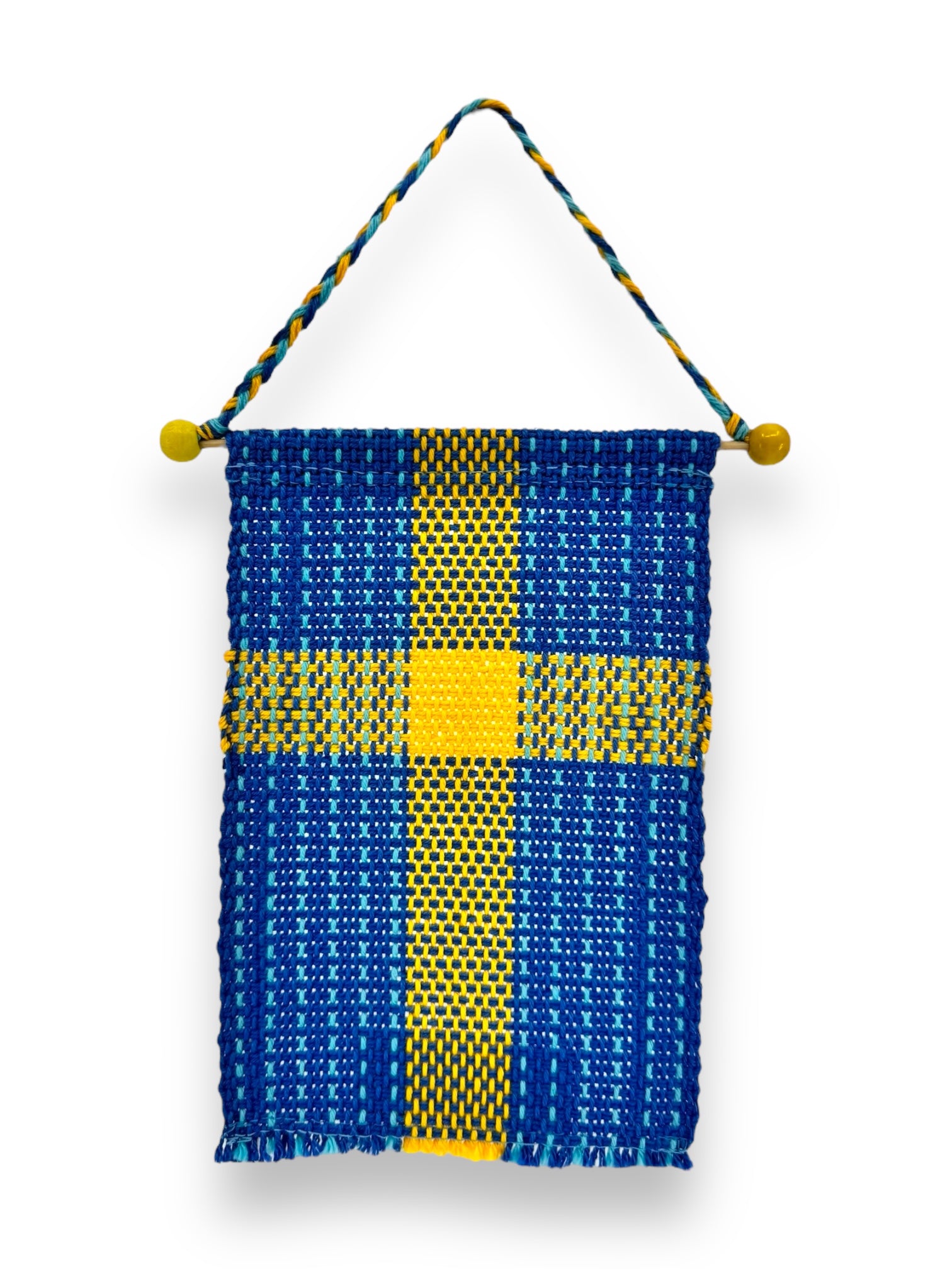 Introducing the Small Swedish Flag on Hanger, a hand-woven tapestry measuring 6.5"x4", featuring a blue and yellow pattern with a central cross reminiscent of the Swedish flag. This piece is crafted from 100% cotton and hangs elegantly from a braided cord with yellow wooden knobs at each end for an authentic touch.