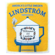 The Dishcloth: Lindström Water Tower features a yellow and blue sign with "Americas Little Sweden, Lindström, Minnesota" and an illustrated teapot water tower labeled "Välkommen till Lindström." Perfect for Swedish-inspired kitchens.