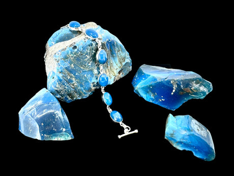 The Plain Oval Seven Stone Swedish Blue Sterling Silver Bracelet, elegantly crafted and featuring seven exquisite stones, is showcased against striking rough blue stones on a black background.