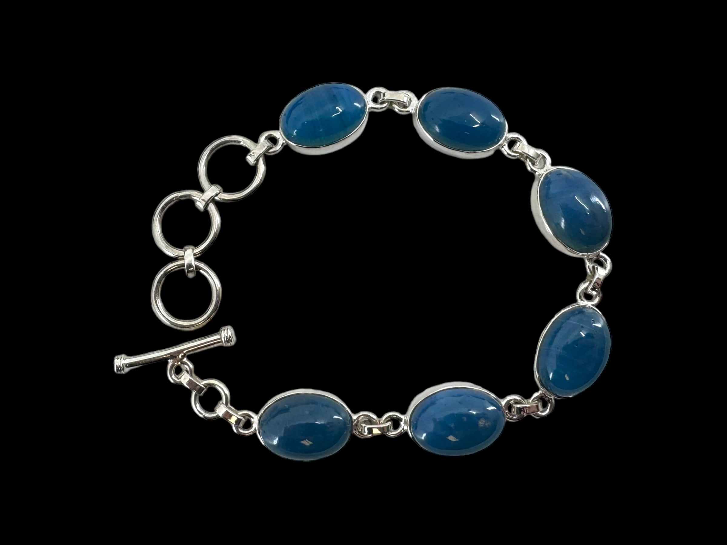 Introducing the "Bracelet: Plain Oval Seven Stone," a sophisticated silver bracelet showcasing a series of linked oval blue stones and completed with a toggle clasp, elegantly set against a black background.