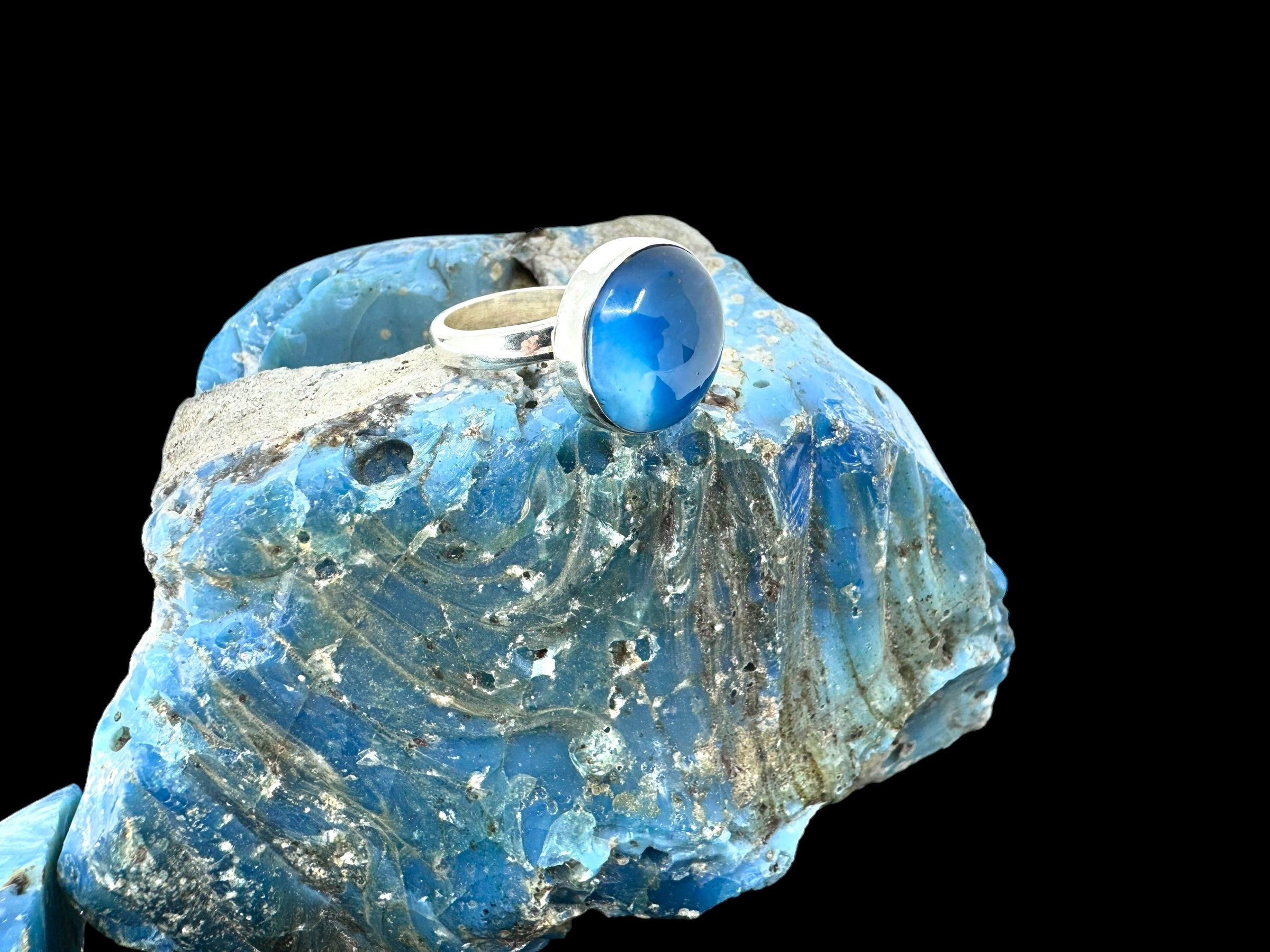 Pain Round - Swedish Blue ring, adorned with a mesmerizing Swedish blue stone, stylishly displayed on a large piece of raw blue mineral set against a black background.