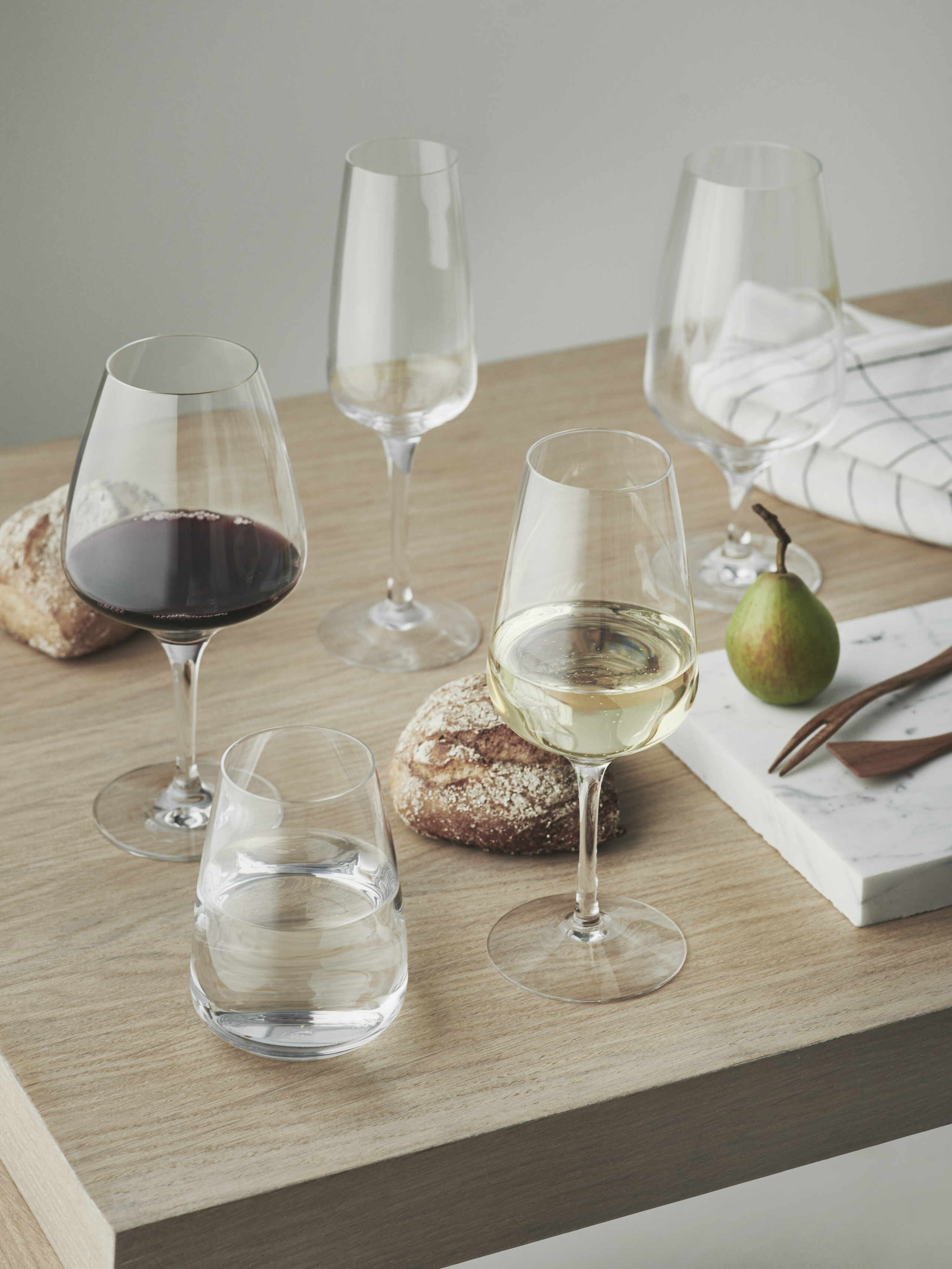 A wooden table elegantly displays four Orrefors Pulse Wine Glasses filled with red and white wine. Crystal glass glistens beside water, bread rolls, a pear, and cutlery on a napkin, all arranged in minimalist Scandinavian design.