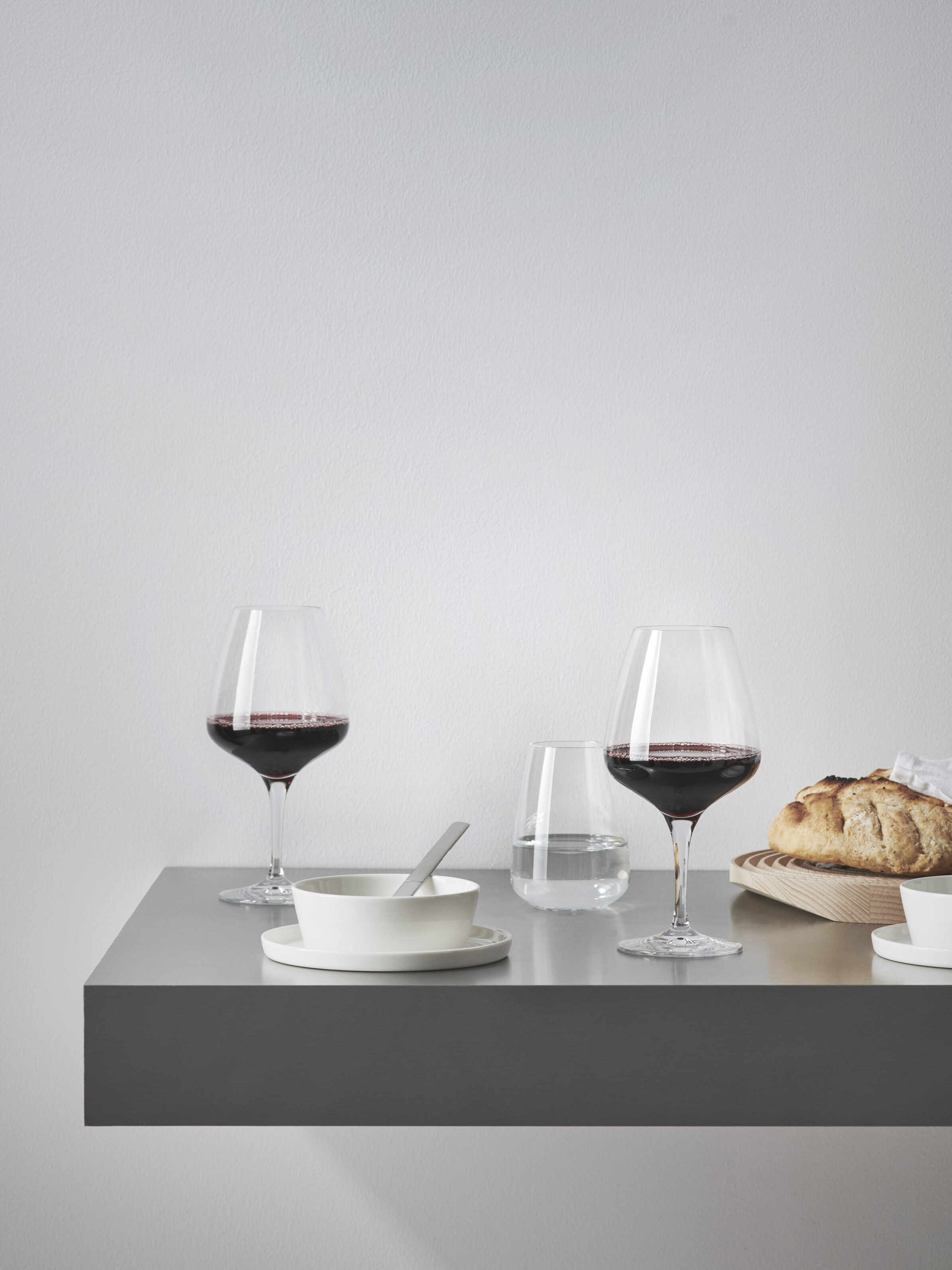 Two glasses of red wine in Orrefors: Pulse Wine Glass 15.5oz, a glass of water, bread, a bowl, and a plate adorn a minimalist gray floating table against a white wall, showcasing Scandinavian design.