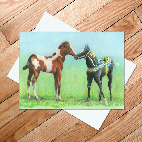 The card titled "Dala Horse Making Friends" features a charming illustration of two painted horses standing on grass, embodying the allure of Swedish Dalahäst design. One horse is brown and white, while the other is black with decorative patterns inspired by Rachael Koppendrayer's style. The image rests against a wooden floor background. The card dimensions are 5x7 inches.