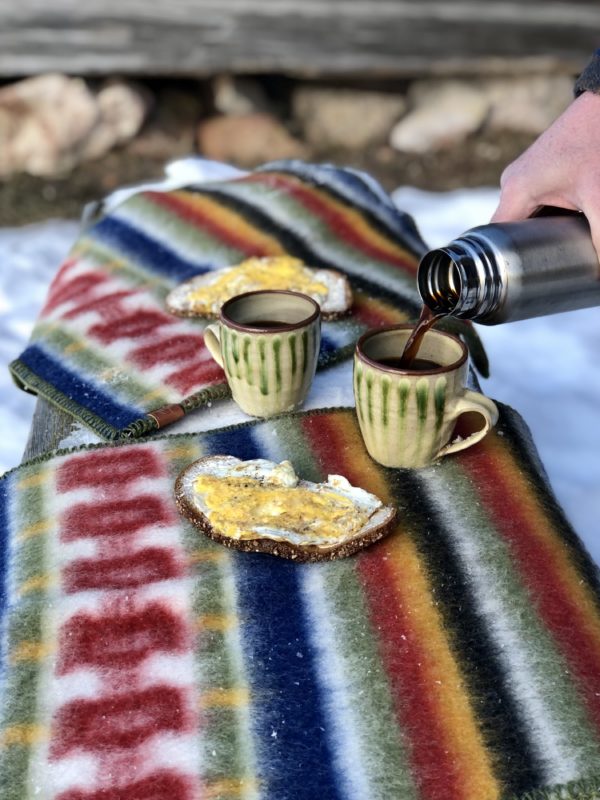 Two ceramic mugs rest on a striped blanket as a hand pours coffee from a thermos. Next to the mugs, there are two slices of bread with spread. The scene is set against snowy rocks and is enhanced by an outdoor sports seat pad made of 100% Norwegian Organic Lambswool in the classic Rättvik pattern by Kerstin Landstrom.