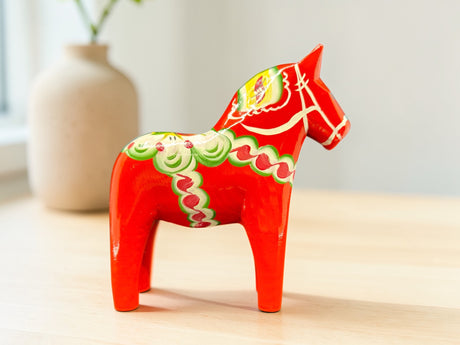The Dala Horse: Red 6" Swedish Dala Horse Figurine is handcrafted in Sweden with green and white patterns, standing on a light wooden surface; a blurred beige vase in the background highlights Nordic folk art tradition.