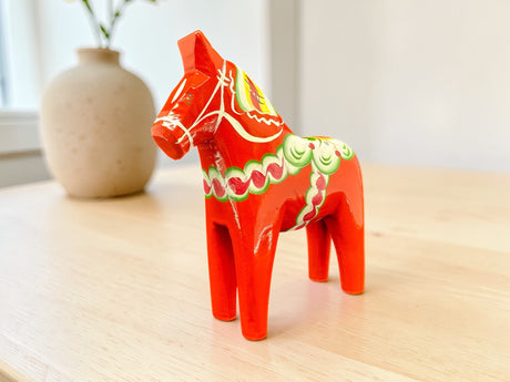 A Dala Horse: Red 6" Swedish Dala Horse Figurine – Handcrafted in Sweden, featuring Nordic folk art motifs, stands on a light wooden table with a beige vase softly blurred in the background.