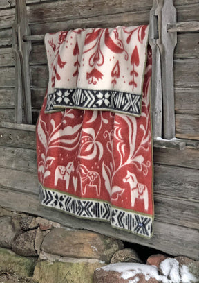 A Blanket: Dalecarlia by Kerstin Landstrom in red, white & black, made of 100% Norwegian lambs wool with Swedish Dala horses, hangs elegantly on a rustic wooden wall.