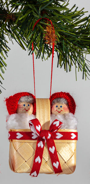 The Swedish Santa Figurines in a Basket is a Christmas ornament showcasing two smiling Santas with red hats, nestled in a basket with a red ribbon and white hearts, elegantly hanging from a pine branch.