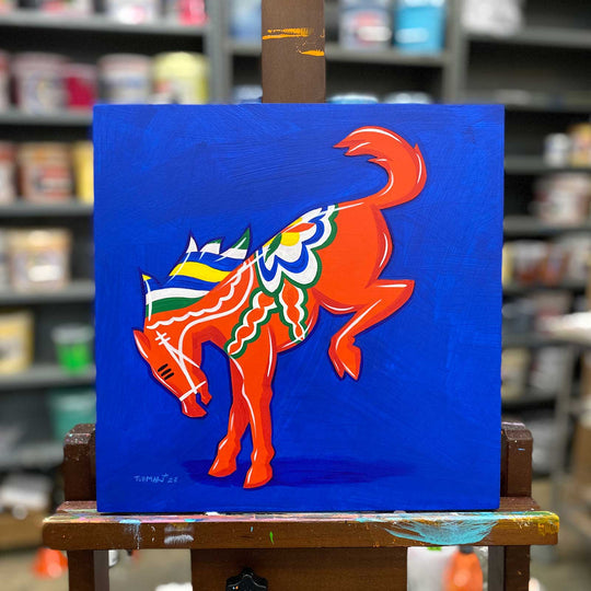 A colorful painting called "Saddle" by Adam Turman, featuring a stylized orange horse reminiscent of a Bucking Dala Bronco, adorned with intricate patterns on a vibrant blue background, is displayed on an easel in an art supply store. The artwork measures 12x12 inches and is framed in black.