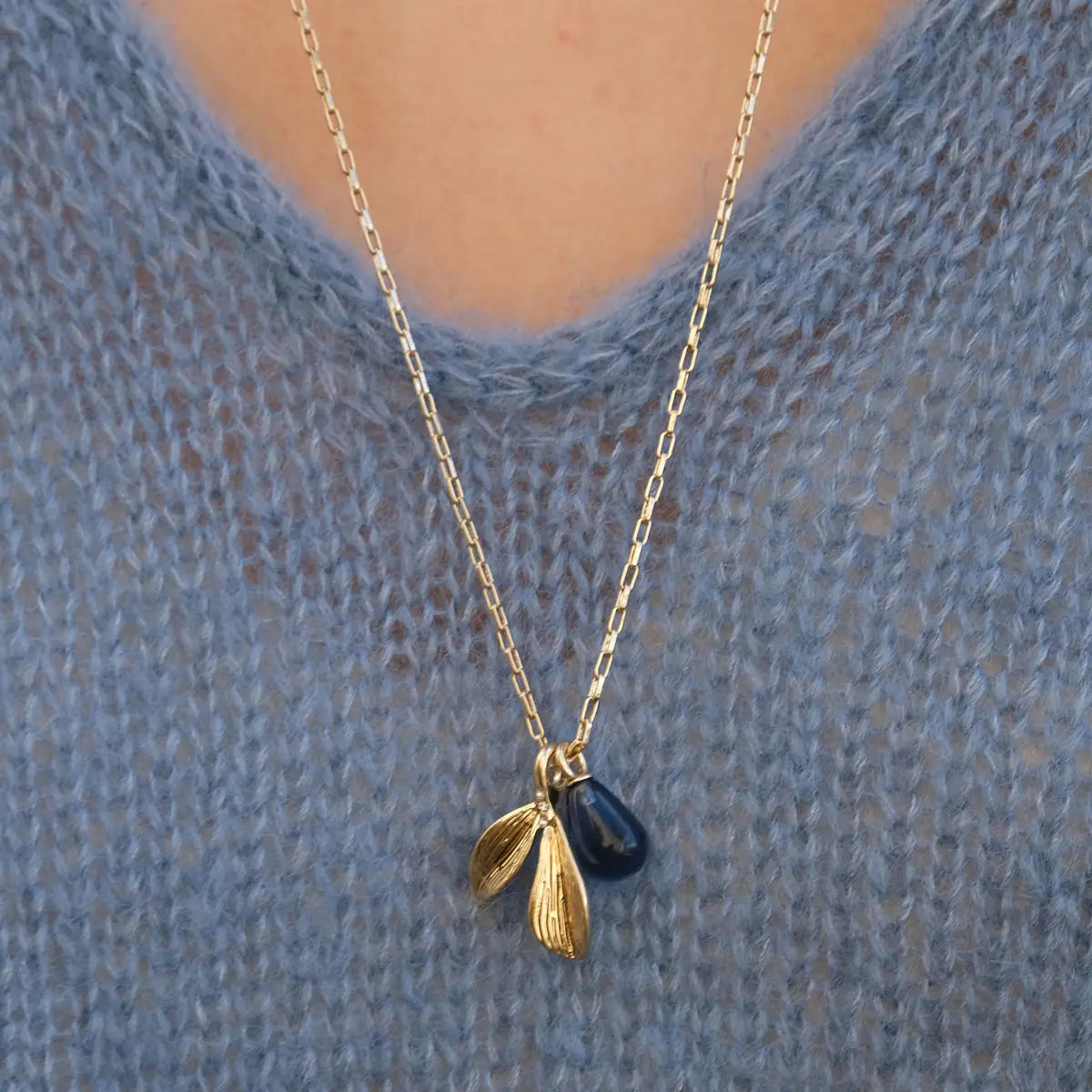 The Necklace: Botanic Hand Cut Glass Blue & Gold features a teardrop gemstone and two gold leaf pendants, beautifully complementing a textured blue sweater.