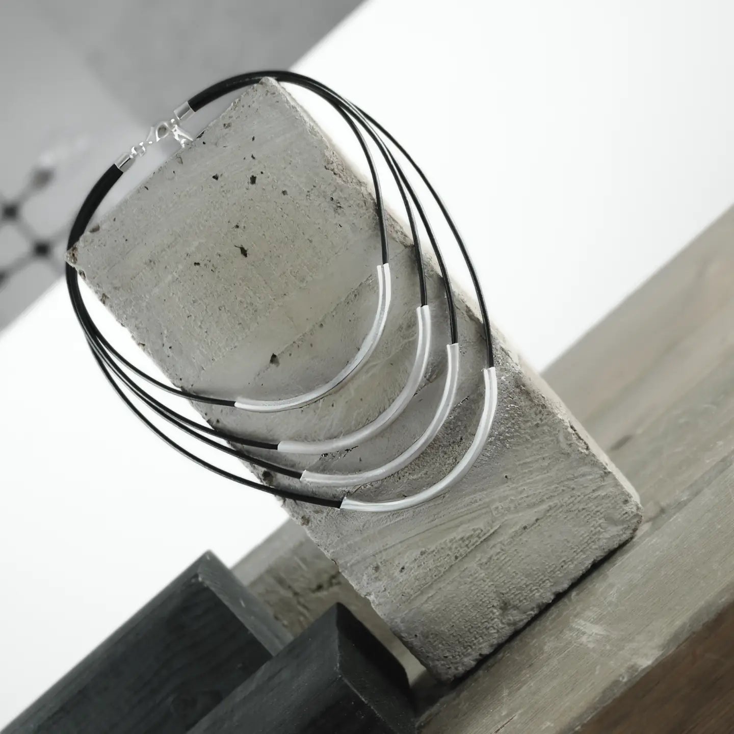 The Necklace: Short Multi String Leather Necklace Silver Plating, featuring silver plating with sleek curved accents, is elegantly displayed on a rectangular concrete block against a gray and white backdrop.