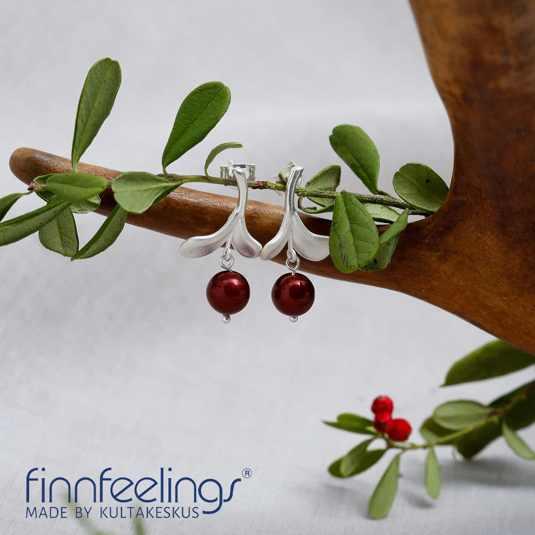 Lingonberry Silver Earrings, adorned with red pearls, are elegantly displayed on a wooden twig with green leaves, embodying Scandinavian design. The words finnfeelings MADE BY KULTAKESKUS highlight the craftsmanship.