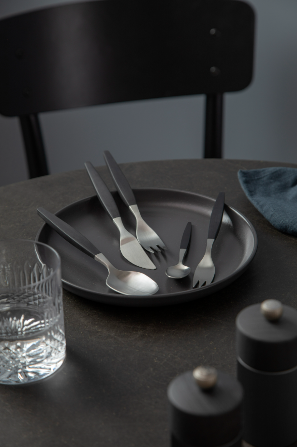 A black plate with Genses Focus De Luxe Cutlery in Black/Matte Steel rests on a dark table alongside a glass of water and a dark napkin, creating a retro classic ambiance. A chair is visible in the background.