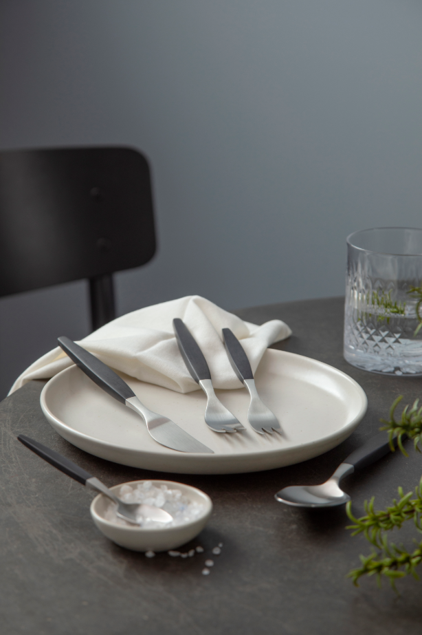 A set of Gense: Focus De Luxe Cutlery in black/matte steel sits elegantly on a white plate with a glass of water, napkin, salt bowl, and spoon on the dark table. Blending modern function with vintage style, this cutlery adds sophistication to any meal setting.