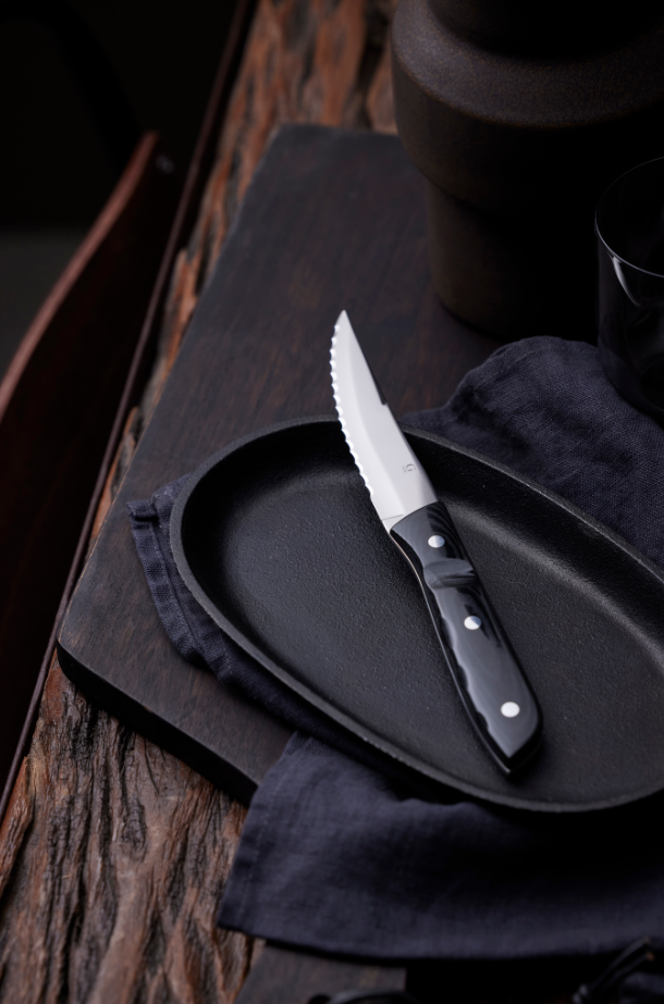 The Gense: Old Farmer Micarta Steak Knife - Steel, featuring a black handle, rests elegantly on a black oval plate atop a wooden table, accompanied by a dark cloth.