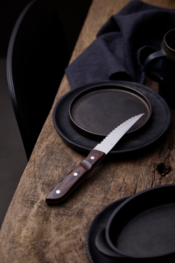 The Gense: Old Farmer Micarta Steak Knife - Steel, with its stainless steel blade and wooden handle, is beautifully placed on an empty black plate atop a wooden table, accompanied by a dark cloth napkin and another black plate.