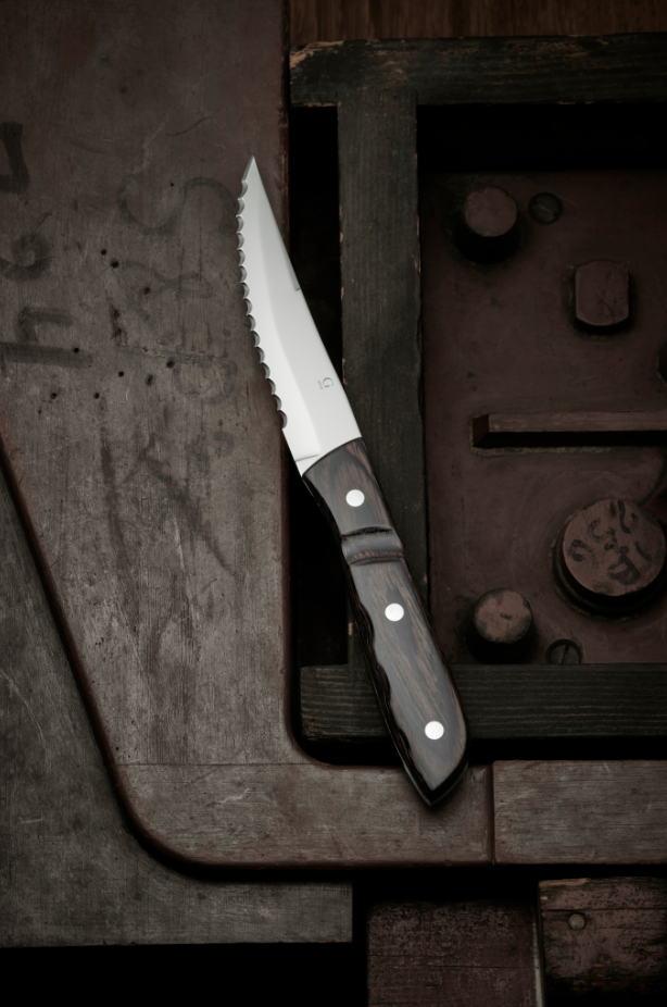 The Gense: Old Farmer Micarta Steak Knife - Steel, featuring a stainless steel serrated blade and dark wooden handle, rests on a rusted metal surface with bolts.