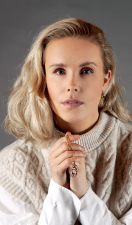 A woman with shoulder-length blonde hair, in a cream cable-knit sweater and white shirt, holds a necklace and looks at the camera, elegantly showcasing the Flor Alida Floral Earrings in sterling silver.