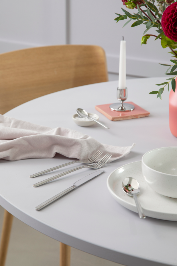 A Nordic round dining table is beautifully arranged with white plates and a Gense: Still Cutlery Set in polished steel, offset by a pink napkin. A single white candle glows beside a vase of red flowers, and an elegant wood chair completes the scene in the background.