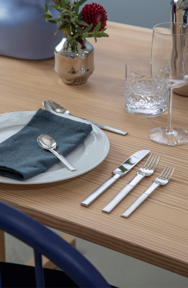 A Swedish-designed dining table features the Nobel Cutlery Set from Gense: A spoon on a gray napkin, meticulously arranged matte/polished stainless steel cutlery, a plate, a glass, and a small vase with red flowers on the wooden surface.