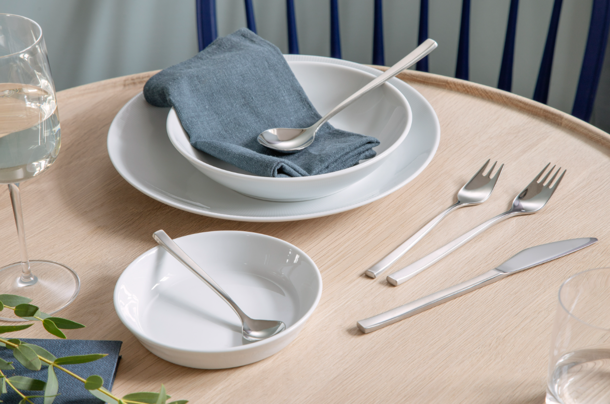 A table setting includes a glass of water, a white bowl with a spoon, and a cloth napkin beside Swedish Cutlery. A side dish and utensils rest on the wooden surface, enhanced by the matte polish of Gense: Fuga Cutlery Set - Matte/Polished Steel. A blue chair and greenery complete the scene.