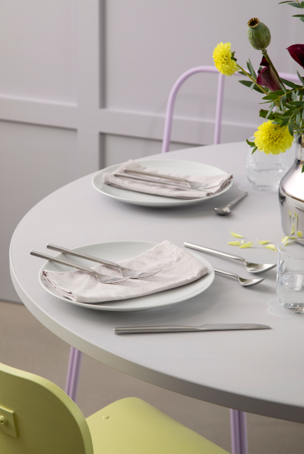 A round table is set with white plates, folded napkins, and Gense: Dorotea Cutlery Set - Matte Steel, complemented by a chic floral arrangement. A light green chair partially visible enhances the modern classic atmosphere.