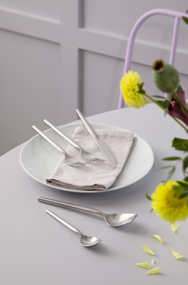 A neatly set dining plate with a folded napkin features the Gense Dorotea Cutlery Set in matte steel, including a fork and knife. Partial views of the table edge and a vase with yellow flowers add to the modern classic charm.