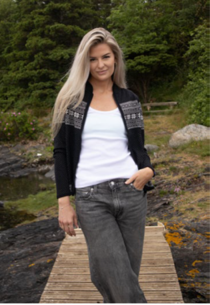 A woman with long blonde hair stands on a wooden dock outdoors, wearing the Northern Story Setesdal Black Norwegian Design Wool Cardigan over a white shirt and gray jeans, with trees and rocks in the background.