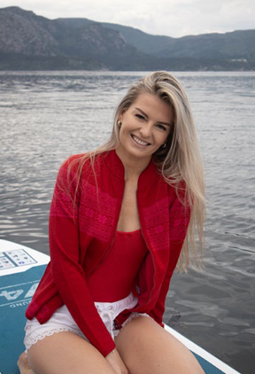 A woman with long blonde hair, in a red top and the Northern Story Setesdal – Red Norwegian Wool Cardigan with white shorts, sits on a paddleboard on a lake with mountains behind her.