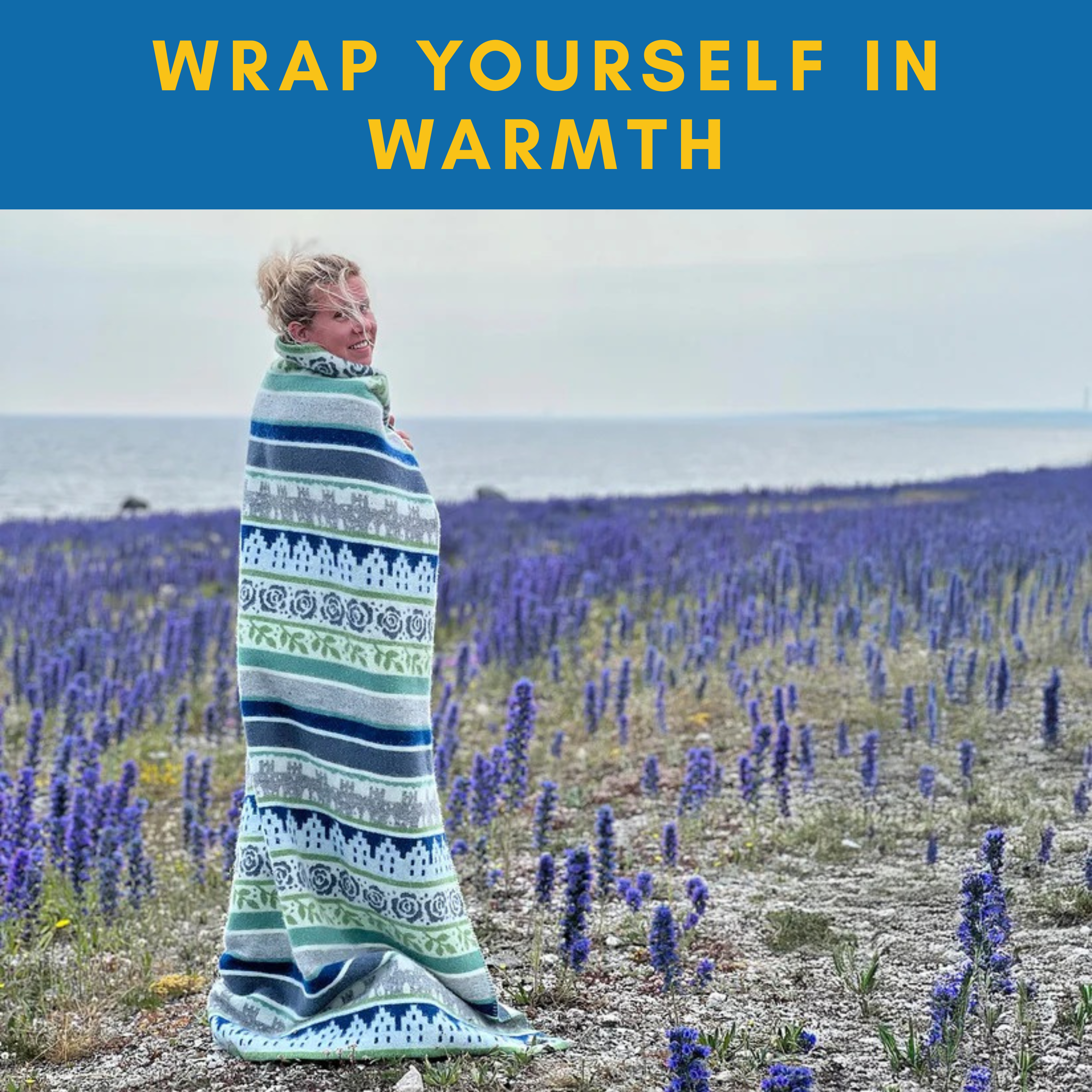 A person wrapped in the Ullfilt Dalarna Wool Blanket—black with Dala horse pattern, 100% Norwegian lambswool—stands in a field of purple flowers by water under a cloudy sky. Text above reads, Wrap yourself in warmth.
