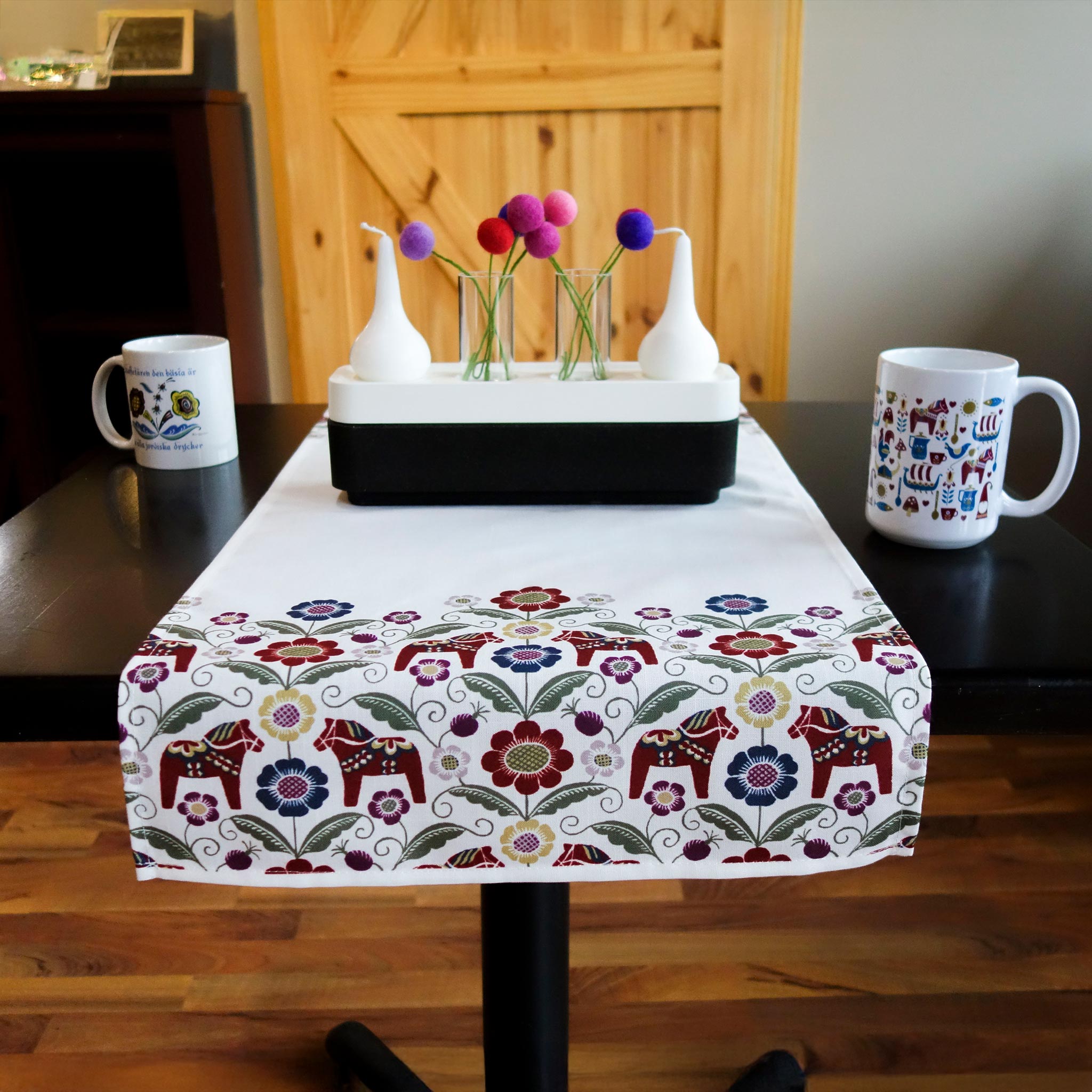 A Siljan Runner - Dala Inspired covers the table with its floral motifs, flanked by two white mugs on each side, and a rectangular vase at the center decorated with colorful circular patterns.