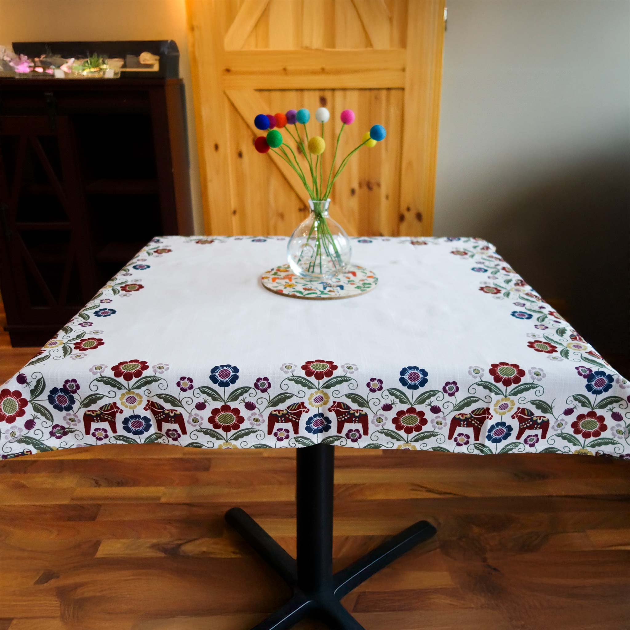 A vase with colorful felt balls sits on a table draped in a Siljan Tablecloth runner, featuring Dala horses and kurbits flowers. The scene is completed by the wooden floor and a charming cabinet beside a wooden door backdrop.