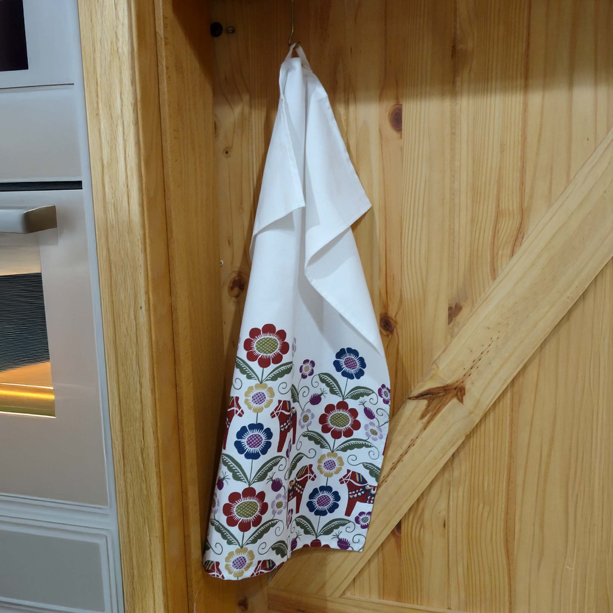 The Siljan Tea Towel, inspired by Dala designs with flowers and kurbits, features colorful floral patterns and charming Dala horses, hanging gracefully on a wooden cabinet.