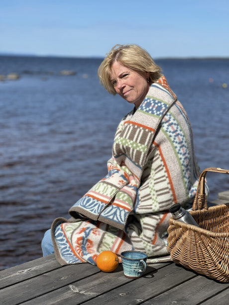 A woman sits on a dock by the water, wrapped in a SKARABORG Wool Blanket (130 × 180 cm), with a basket, an orange, and a mug beside her beneath a clear sky.