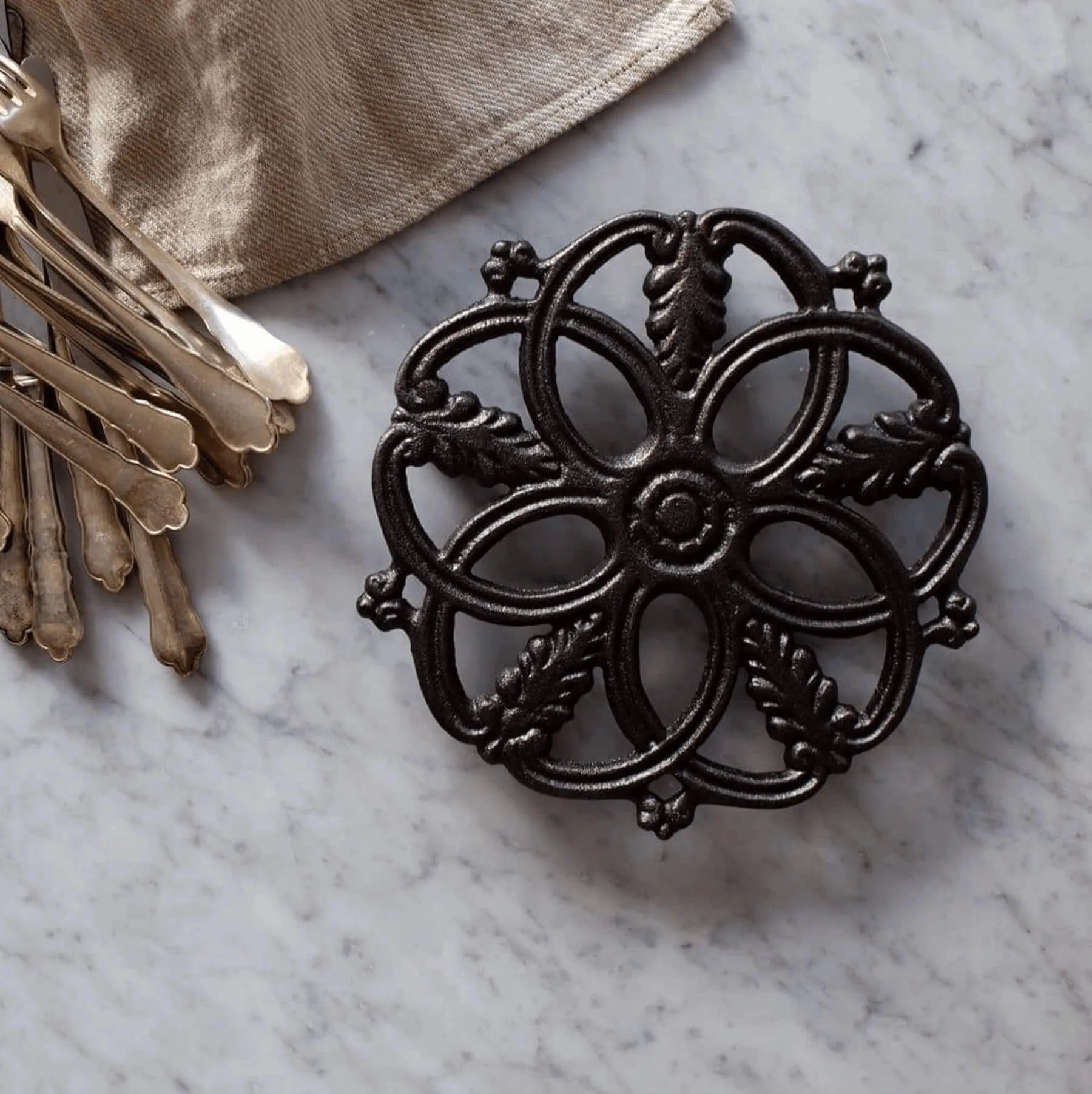 A Skeppshult: Cast Iron Trivet, featuring an ornate floral design and handcrafted in Sweden, elegantly sits on a marble surface beside vintage silver forks.