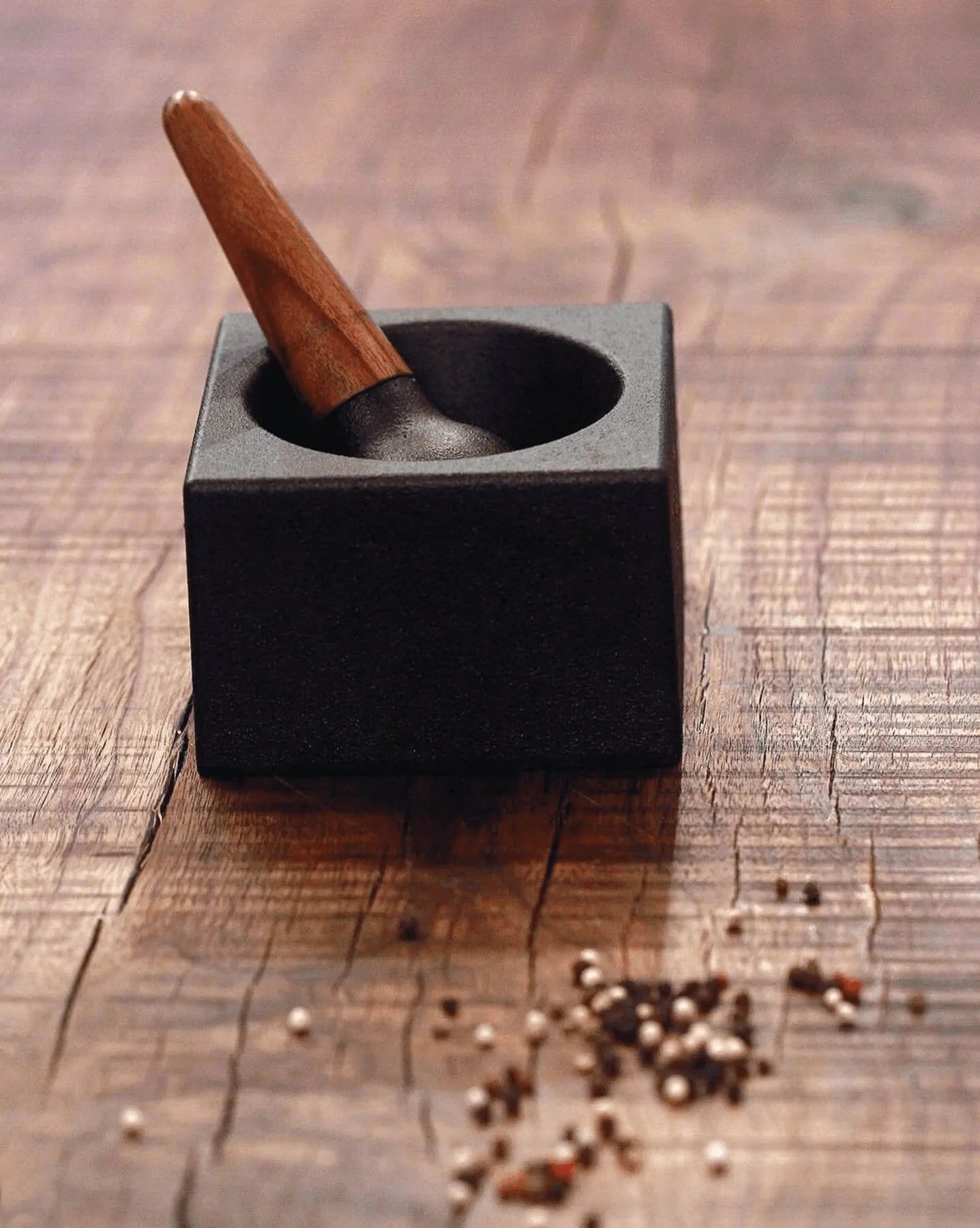 The Skeppshult: Mortar With Walnut Pestle, featuring a square black cast iron design and walnut pestle, sits on a wooden surface with a small pile of mixed peppercorns nearby.