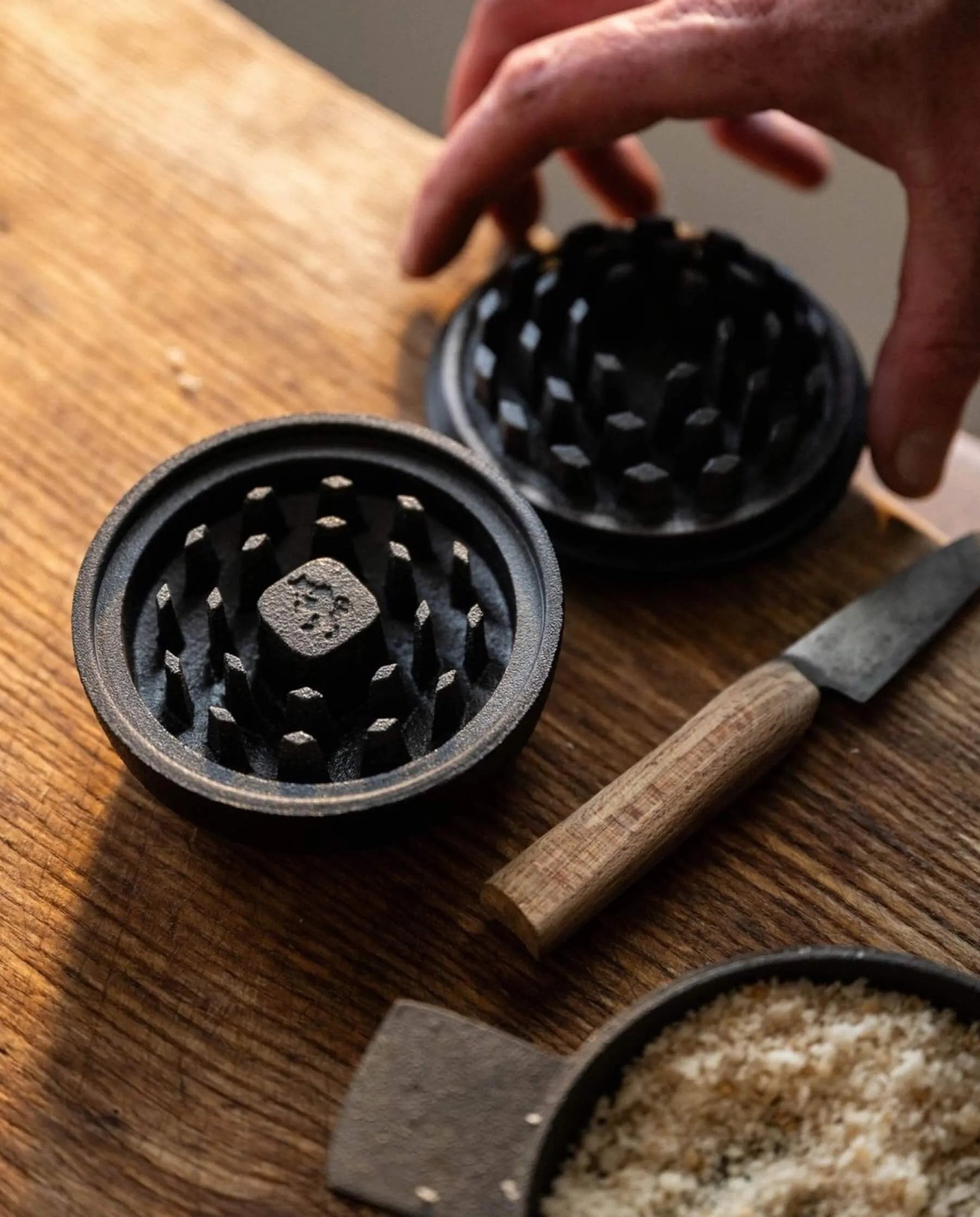 A hand reaches for a Skeppshult: Spice Grinder/Crusher on a wooden surface, its cast iron gleaming beside a knife and grated substance.