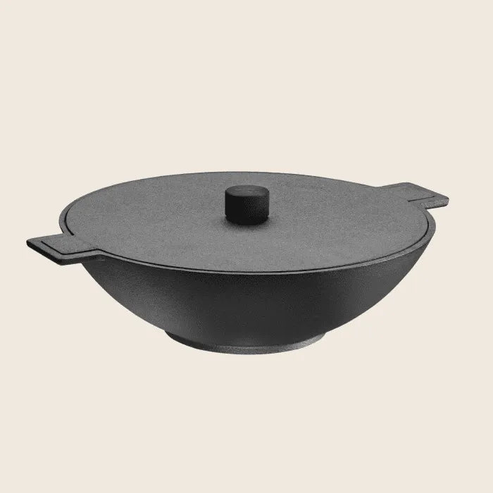 The Skeppshult Wok 13.25 (34 cm), made from premium black cast iron, features a flat lid and dual side handles. Set against a plain white backdrop, it highlights its durable build and non-stick surface, ideal for cooking enthusiasts.