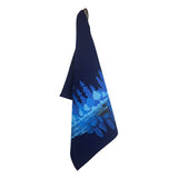 The Midnight Loon Tea Towel, featuring a printed design of a blue river and pine trees, hangs on a hook, capturing the serene essence of Minnesota Midnight.
