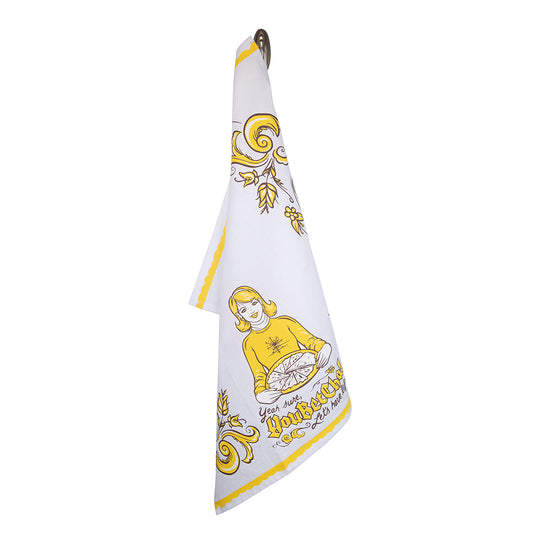 The "You Betcha" tea towel, crafted from white and yellow cotton, features a vintage-style illustration of a woman holding a turkey amid decorative floral patterns. Made in the USA and machine washable, it seamlessly combines charm with practicality as it hangs on a hook.