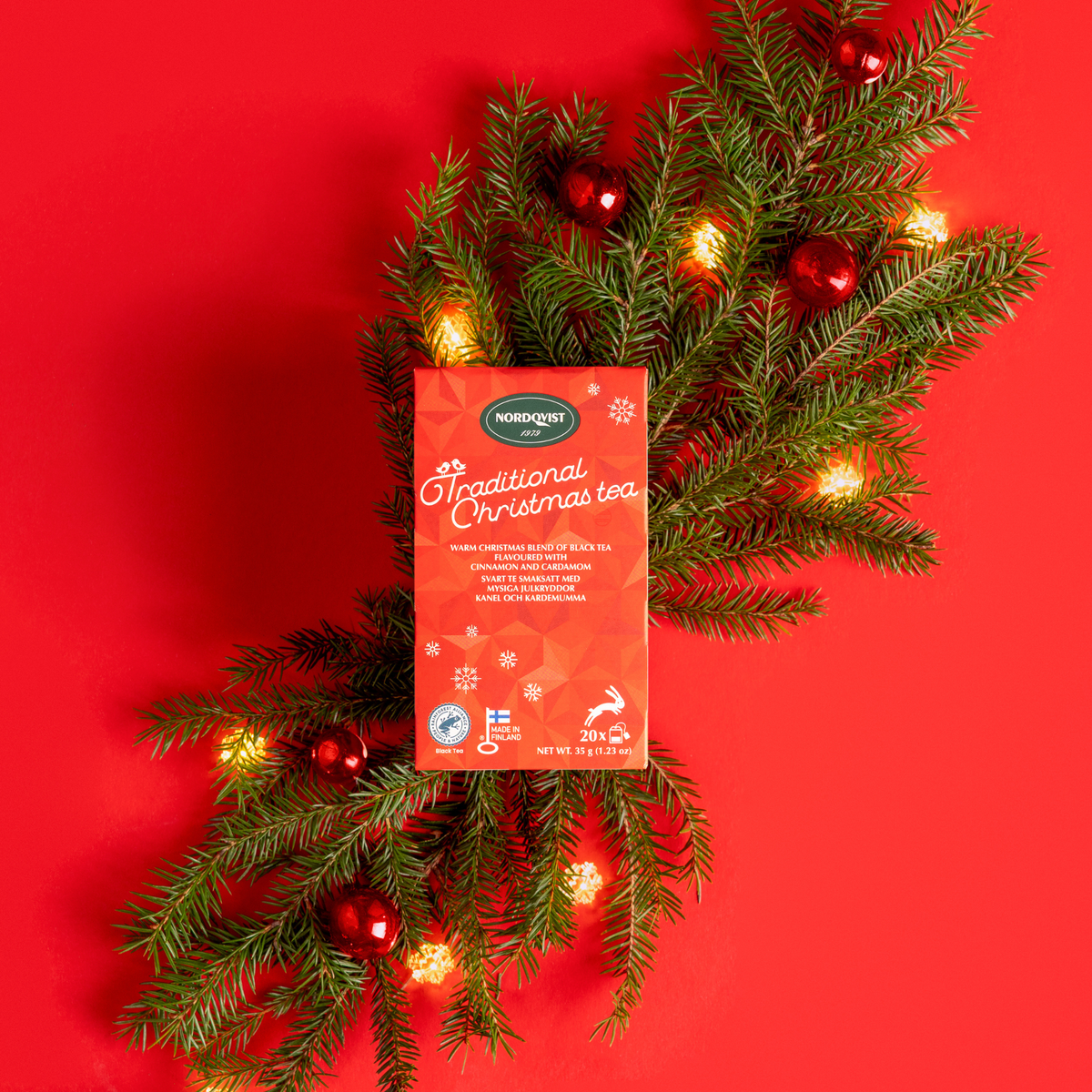 Box of Finnish Christmas black tea set on pine branches with red ornaments and string lights against a red background.