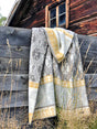 A Brunkulla Cornflower Brown blanket by Kerstin Landstrom, crafted from 100% Norwegian lambs wool, featuring grey and yellow designs, drapes over a wooden fence with an old wooden building in the background.
