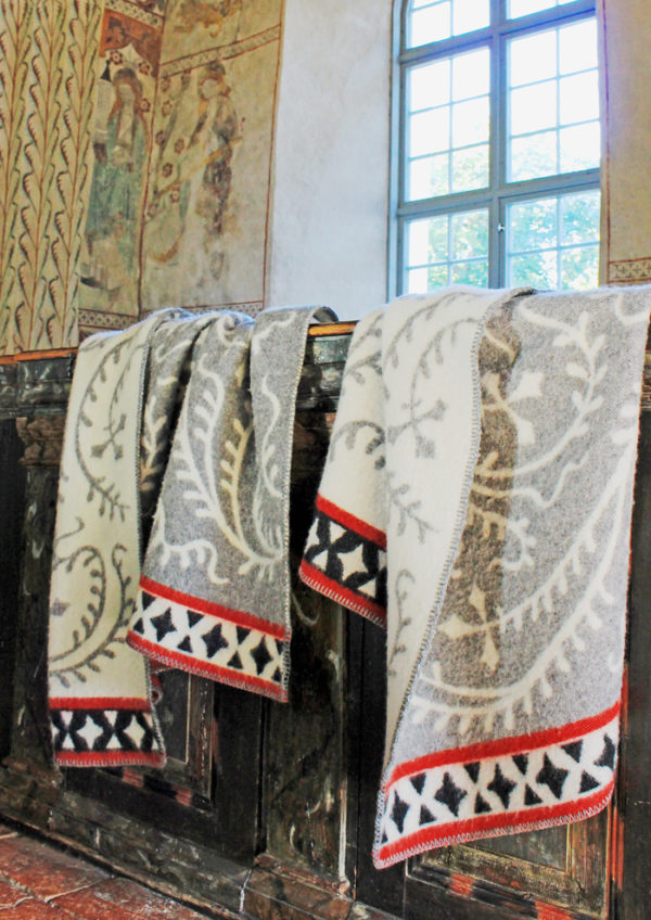 Four Albertus blankets, crafted from 100% Norwegian lambs wool and OEKO-TEX-certified materials by Kerstin Landstrom, elegantly drape over a dark wooden railing. They add cozy charm to the room with painted walls and a large window inviting natural light.