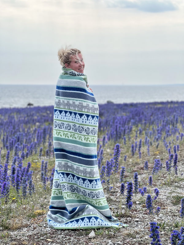 Wrapped in a Wool Blanket: Visby, Blue/Grey, a person stands in a field of purple flowers by the sea under cloudy skies, capturing the essence of Swedish scenery.