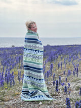 Wrapped in a Wool Blanket: Visby, Blue/Grey, a person stands in a field of purple flowers by the sea under cloudy skies, capturing the essence of Swedish scenery.