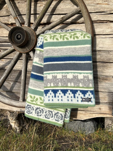 A Wool Blanket: Visby, Blue/Grey with bright stripes elegantly hangs on a wagon wheel, capturing Swedish charm. Nearby, a small cat sits contentedly.