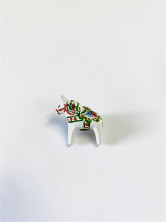 The "Pin: Dala Horse" is a delicate small white ceramic figurine depicting a horse, adorned with green and red painted details that reflect Swedish heritage, all set against a plain white background.